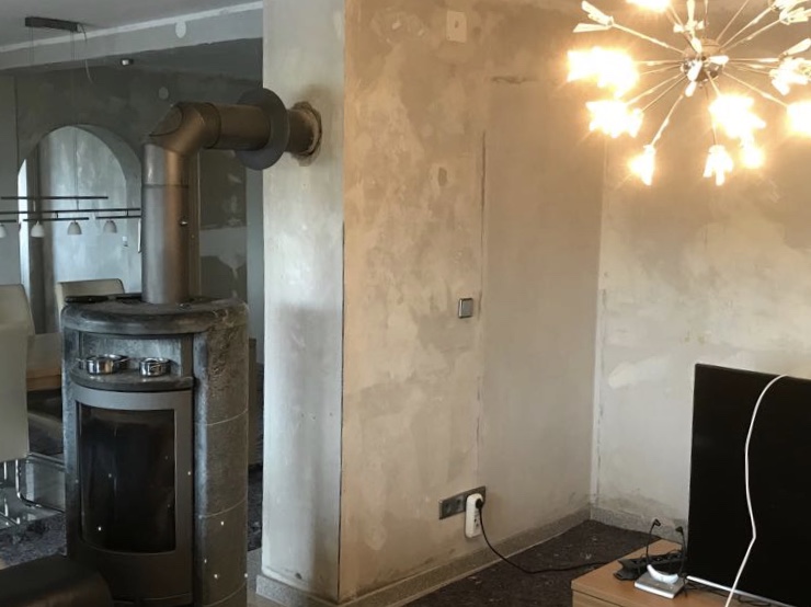 Ein Raum mit einer Holzofenheizung, einer Wand mit unvollendetem Putz, einem Fernseher auf einem Tisch und einer Kronleuchterbeleuchtung.