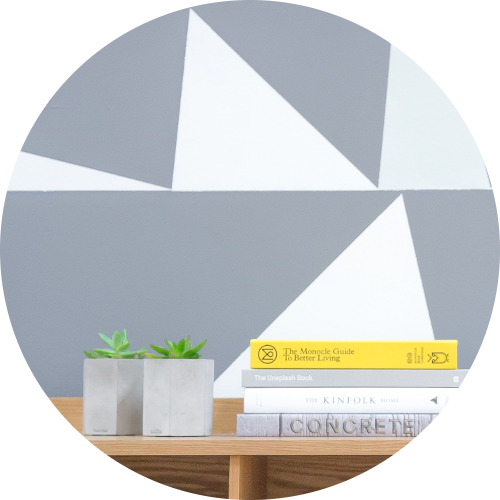 Ein runder Ausschnitt zeigt ein minimalistisches Interieur mit geometrischen Dreiecken an der Wand, einem Holztisch mit zwei Pflanzenkästen und drei aufgestapelten Büchern.